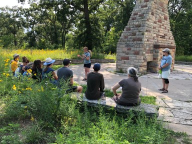 2 teachers standing in front of students seated on a bench in the outdoors.